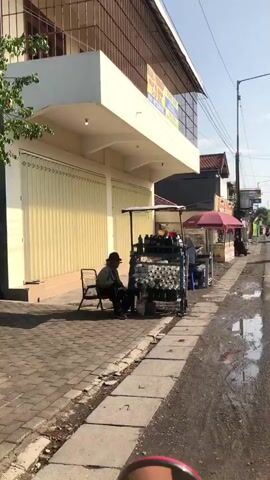 The video shows a street scene with several vendors. In the foreground, on the left, a person wearing a hat and a jacket sits on a chair next to a cart filled with various items. In the middle ground, a person wearing a hooded jacket and jeans sits at a table under a large red umbrella, appearing to be working on something. To the right of this table, another vendor operates under a similar red umbrella. A motorbike is parked nearby. The environment appears to be a commercial street with buildings lining one side and a road with puddles on the other. The overall impression is one of everyday street commerce.