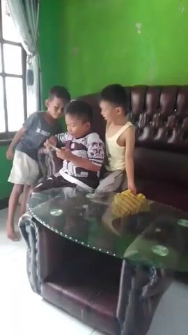 The video shows three young boys in a room. The room has green walls and a brown leather couch. In the foreground, there is a glass-topped coffee table. One boy, wearing a gray t-shirt and white shorts, stands on the left, looking towards the center. Another boy, wearing a patterned shirt, sits on the couch in the middle, holding a mobile phone. A third boy, wearing a yellow tank top and gray pants, kneels on the couch to the right, facing the center. He is holding a yellow object, possibly a piece of cloth, which he is waving. The boy in the middle appears to be showing something on his phone to the other two boys. The boy on the right is leaning forward, looking at the phone, and the boy on the left is also looking at the phone. The boy on the right raises his arms and then lowers them. The video appears to be a recording of everyday life.