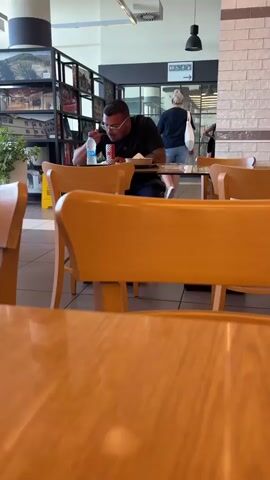 A man wearing a black t-shirt and glasses is sitting at a table in a food court, eating. He is holding a can of soda and a bottle of water. In the background, other people are walking around and sitting at tables. The food court appears to be clean and well-lit.