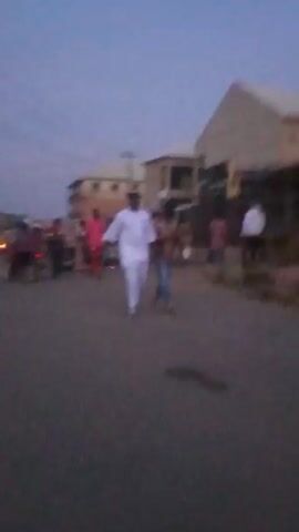 The video shows a street scene at dusk. There are several people walking and a few motorcycles with their lights on. One person is wearing a white outfit. The footage is blurry. The background includes buildings and power lines.