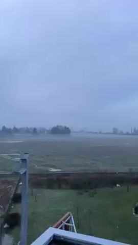 The video shows a landscape on a foggy day. In the foreground, there is a green lawn and a glimpse of a building structure. Beyond that, a field is partially obscured by fog, with hints of vegetation visible. A dark line, possibly a road or track, cuts across the field. In the distance, faint silhouettes of trees and building structures are barely discernible through the fog. As the camera pans to the right, modern, multi-story buildings with brown and white paneling come into view, dominating the foreground. The sky is overcast, contributing to the muted, somber atmosphere.