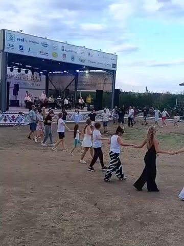 A group of people are dancing in a circle in front of a stage. The stage has a banner across the top with logos and text. There are musicians on the stage and people watching. The ground is dirt and there are some trees and bushes in the background. The sky is cloudy. The people are holding hands and moving in a circular motion. Some of the people are children and some are adults. The adults are wearing casual clothing. The children are wearing dresses and shorts. The overall atmosphere appears to be festive.