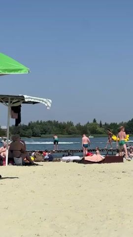 This media file captures a lively summer day at a beach in Verbizh, Ukraine. The scene is set on a sandy shore bordering a calm body of water, likely a lake or river, with lush green trees lining the far bank. The sky is a clear, vibrant blue, indicating fair weather and a sunny day, likely in the late morning or early afternoon given the bright sunlight and shadows.

Several people are present, enjoying the water and the beach. In the foreground, a large green umbrella shades a few individuals relaxing on lounge chairs, with one person seated and facing away from the camera. Beach bags and towels are scattered around.

Further out, a wooden pier extends into the water, with several people standing and walking on it. One person is positioned at the end of the pier, seemingly about to jump or dive into the water. Nearby, a bright yellow inflatable object, possibly a towable water toy, is being pulled across the water by an unseen boat, creating a wake. More figures are visible on the pier and in the water, some appearing to be swimming or wading.

The overall atmosphere is one of leisure and recreation. The people appear relaxed and engaged in summer activities. The presence of the pier, the inflatable water toy, and the clear weather all contribute to a typical beach day scene. No specific text is clearly visible or legible in the provided frames.