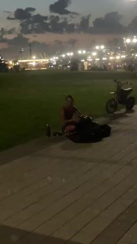 At dusk, a person is sitting on the ground next to a black bag. A black water bottle is to their left. A motorcycle is parked to their right. In the background, lights are visible from buildings and streetlights. The sky is dusky with clouds. Two people walk by in the foreground. The person sitting on the ground appears to be interacting with the black bag. The video is shaky.