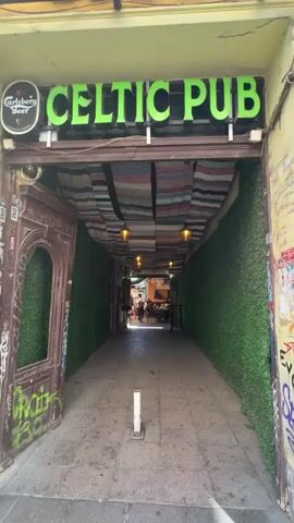 The video shows a narrow alleyway leading to a pub called "CELTIC PUB." The alleyway is lined with green artificial grass on both sides. Colorful woven blankets are draped across the ceiling of the alley, with lamps hanging from them. Graffiti is visible on the left side of the entrance and on a wooden door. A sign for "Carlsberg Beer" is visible above the pub's entrance. In the distance, at the end of the alley, people are seated at tables outside what appears to be a seating area, and there are flags visible on a wall. One person is walking down the alley towards the viewer. The ground is paved.