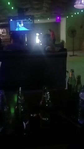 The video shows the interior of a dimly lit room with a TV screen in the background, showing a scene with a person walking on a white surface. In the foreground, there's a table with several empty beer bottles and a water bottle. The room appears to be a bar or lounge area, with chairs and a fan visible. The lighting is varied, with green fairy lights, a purple lamp, and the glow from the TV screen. The camera moves slightly, changing the perspective and focus. There's a person sitting on a chair in front of the TV. The room seems to be decorated with rough, textured walls. Background noise suggests music or chatter.