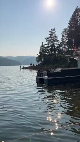 The video shows a body of water, likely a lake, with trees along the shoreline. The sun is shining brightly in the sky, creating reflections on the water's surface. A boat with a dark hull and white superstructure is moored near the shore. There are people on the shore and on the boat. One person is standing on the shore, and a group of people is gathered further down the shore. Another boat, a catamaran, is visible on the right side of the frame, with two people on board. One person is wearing an orange life jacket and sitting on the deck of the catamaran. The scene suggests a recreational activity on the water, possibly swimming or boating. The weather appears to be clear and sunny.
