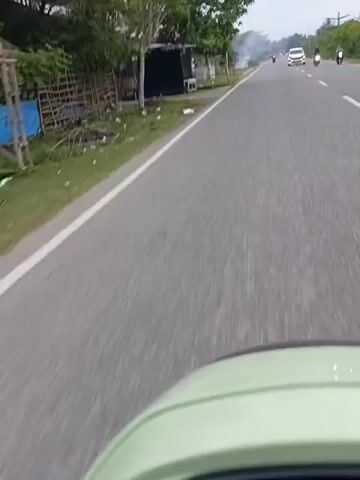 The video depicts a first-person view from a vehicle, likely a scooter or motorcycle, traveling on a paved road. On the left side of the road, there is green grass with a small, simple structure, and further back, there are trees. On the other side of the road, traffic includes a white car and at least one motorcycle moving in the same direction. There is a plume of smoke rising in the distance. The view is partially obstructed by the top of the vehicle's dashboard, which is a light green color.