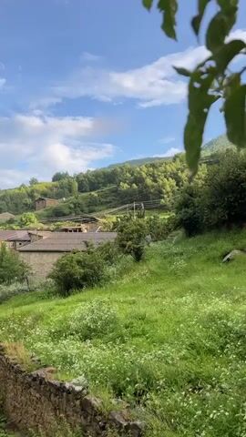The video shows a wide shot of a village nestled in a green, hilly landscape under a blue sky with scattered clouds. In the foreground, there is a low, crumbling stone wall partially covered in grass. A grassy slope rises behind the wall, dotted with small white wildflowers. Several stone structures, possibly ruins, are visible along the bottom edge of the frame.

As the video progresses, the camera pans slightly to the left and upward, revealing more of the village. Houses with tiled roofs are scattered across the hillside, surrounded by trees and vegetation. Power lines are visible on poles running through the village.

In the latter half of the video, a vibrant rainbow arcs across the sky to the left, with its end touching the distant mountains. A few scattered trees with green foliage are in the mid-ground. On the lower part of the hill, there are two cows grazing in the green grass. The overall impression is of a rural, peaceful setting with a touch of natural beauty, enhanced by the appearance of the rainbow.