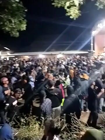 This is a video of a large gathering of people outdoors at night. The crowd appears to be watching a performance on a stage, as evidenced by the lighting and a band visible on the stage. There are trees and foliage in the foreground, and some structures in the background, suggesting an open area like a field or a venue. The people are dressed in casual clothing, and the atmosphere seems lively. The video is shaky at times, indicating it was likely filmed by someone in the crowd.