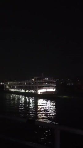 At night, a brightly lit passenger ship rests on the water. The ship is multi-tiered with many arched windows and lights illuminating its form. The surrounding water reflects the ship's lights, creating shimmering patterns. The night sky is dark, adding to the contrast of the illuminated vessel against its background. A railing appears in the foreground, likely indicating the vantage point of the viewer. The setting appears to be a river or harbor.
