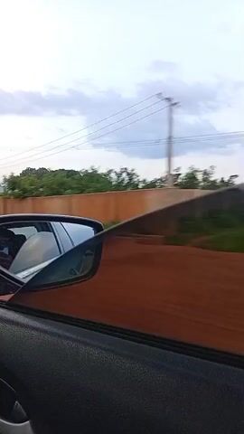 The video shows a view from inside a car looking out the passenger window as the car travels along a road. A tinted window is partially rolled down. In the side mirror, a reflection of the interior of the car can be seen, including a person. Outside the car, there is a brown wall, some trees, houses, and utility poles with wires against a cloudy sky. The scene appears to be a residential area.