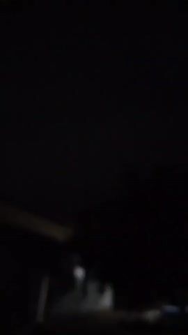 A dark video shows an exterior view of a building at night. The camera is shaky, and the lighting is poor, making it difficult to discern specific details. A window on the building appears to be lit from within. There are trees or other foliage visible in the foreground, also obscured by darkness. The overall impression is of a low-light, possibly handheld recording of an outdoor space.