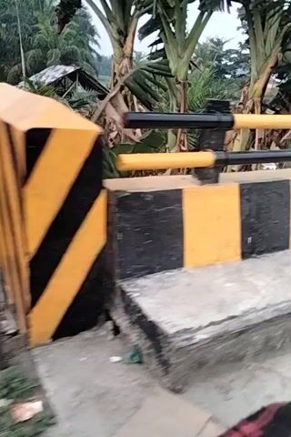 The video shows a view from a moving vehicle traveling alongside a street. The street is lined with a concrete barrier painted with alternating yellow and black stripes. Beyond the barrier, there is a body of water with several small boats and structures on the opposite bank. Trees and some buildings are visible in the background. The scene appears to be a rural or semi-urban environment. The camera is angled downwards and to the side, with a slight blur indicating movement.