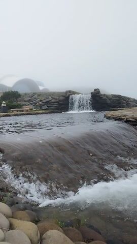 This is a vertical, close-up video of a small waterfall. The water is clear and flows over rocks. The surroundings appear to be natural, with rocks and vegetation visible. The sky is overcast, creating a diffused light.