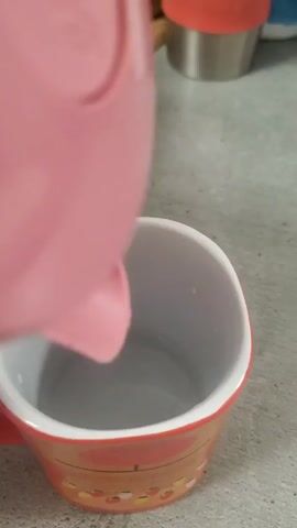 A pink kettle is pouring water into a mug with an orange and white pattern. The mug is on a gray surface. In the background, there is a silver container with an orange lid.