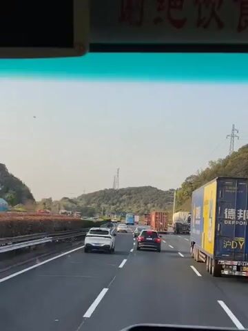 The video shows a view from inside a vehicle, looking out onto a busy highway. Several lanes of traffic are visible, including cars and large trucks. A blue and yellow "DEPPON" truck is prominently seen in the right lane. The highway is surrounded by green, hilly terrain under a clear sky. The sign above the windshield has Chinese characters. Traffic appears congested with vehicles moving at slower speeds. No people are visible inside the vehicle, only the road and other vehicles ahead.