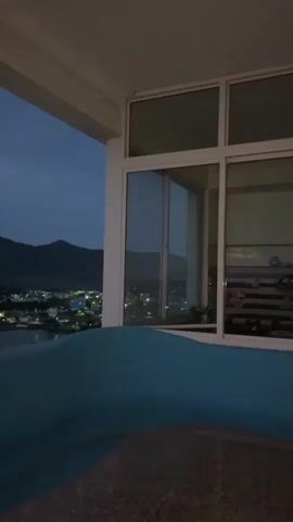 The video shows a view from a balcony at dusk. In the foreground, a blue cushioned seating area is visible. Beyond the balcony, a city lights up as night falls. Mountains can be seen in the distance. The sky is a deep blue with scattered clouds. The overall atmosphere is peaceful and serene.