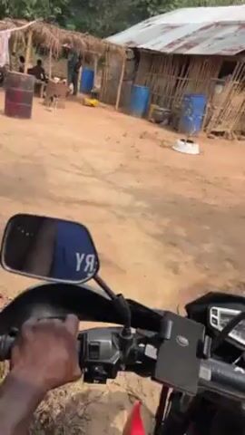 The video begins from the perspective of someone sitting on a motorcycle, facing a yard. In the yard there is a group of people sitting or standing under a thatched roof structure. To the right of this is a building constructed of sticks with a metal roof. There are several blue plastic drums scattered around the yard. A white dog is also visible. The camera moves forward, showing a dirt yard with green leaves scattered on the ground and several structures made of sticks. There are trees and green vegetation in the background.