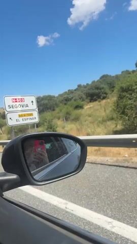 The video shows a view from the passenger side of a car driving on a road on a sunny day. The sky is blue with a few white clouds. A green hill with dry grass and trees is visible on the right side of the road. A guardrail runs along the side of the road. In the rearview mirror, a person holding a red phone is reflected. A white triangular road sign with a snowflake symbol and the text "3 km" is visible on a pole to the right of the road. Another circular sign with the number "40" inside a red circle is seen on the left side of the road in the distance. The car appears to be moving at a moderate speed.