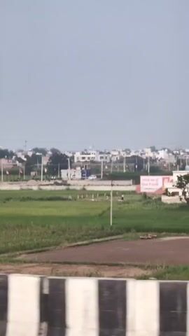 The video captures a view of a rural landscape under a cloudy sky. In the foreground, there is a concrete barrier with black and white stripes. Beyond the barrier, a green field stretches into the distance. Further back, a cluster of houses and buildings can be seen. One prominent building is white with a few windows. To the left of this building, a large billboard with an image and text is visible. In the middle ground, there are more green fields, some of which appear to be cultivated. In the background, trees and the outlines of more buildings and infrastructure, including power lines, are discernible against the hazy sky. A single person is visible walking in the far left of the frame, near the billboard.