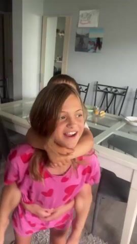 Two young children are the subjects in this video clip. The older child, a girl with blonde hair, is wearing a pink top and shorts with red hearts. She is carrying a younger child, a boy, on her back. The boy is wearing a white t-shirt and appears to be hugging the girl. The children are in an indoor setting that appears to be a dining or living area. A white dining table with a glass top is visible in the background, along with chairs. There are also some framed pictures on the wall and a mirror. The lighting suggests it is daytime. The children seem to be playing or interacting playfully. There is no visible text in the video.