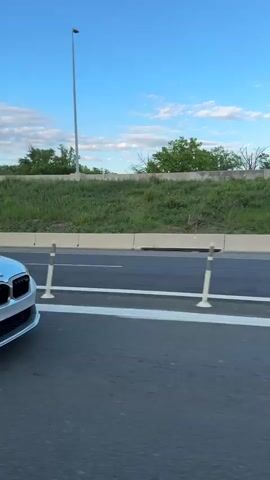 A silver BMW M5 F90 is seen from another vehicle on a highway. The BMW is on the right and passes the camera car. The BMW accelerates and passes other cars. The sky is blue with some clouds. The highway has green grassy areas on either side with a line of trees. Light posts line the road. The BMW has a license plate that reads CS F90. Other traffic is visible in the distance.