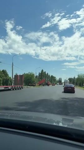 A daytime video taken from inside a moving vehicle shows a road in Pervomaiskoye, Russia, under a bright blue sky with scattered white clouds. The scene suggests ongoing road construction or maintenance. On the left side of the road, a large flatbed truck with a tall, bulky load is parked. Further ahead, several orange or red dump trucks are visible, with one having its bed raised, indicating dumping activity. Two road rollers are also present, suggesting paving or compaction work.

Cars are driving on the road, navigating past the construction equipment. A red sedan is seen ahead of the camera car, and a white tanker-like truck is further down the road. Trees line both sides of the road, and streetlights are visible. A traffic light can be seen in the distance. No specific text is legible on any signs or vehicles.