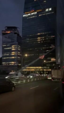 The video captures a night-time scene from a moving vehicle, likely driving on a multi-lane road or bridge in an urban environment. Towering skyscrapers dominate the background, their windows glowing with interior lights, indicating active businesses or residences. The sky is dark, suggesting it's evening or night.

In the foreground, a black minivan drives on the left, while other cars and trucks with illuminated headlights and taillights are visible in the distance. Streetlights cast light onto the road.

A prominent feature is a large glass skyscraper on the right, displaying "GROUPE BPCE" in illuminated letters near its top. Another, slightly smaller building to its left also shows some illuminated details and a logo that appears to be "RX" or similar. The general atmosphere is that of a bustling city after dark, with artificial lights illuminating the urban landscape. The provided context indicates the location is Ivry-sur-Seine, France.