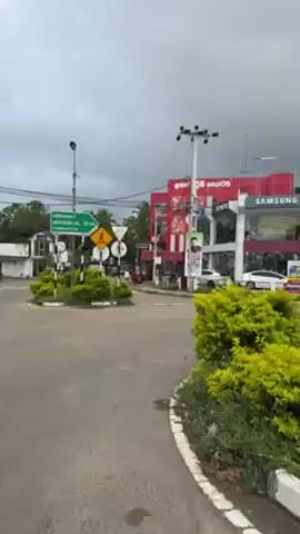 The video shows a roundabout in a Sri Lankan town. The sky is overcast. Several businesses, including Singer, Samsung, and Panasonic, are visible around the roundabout. There are also traffic signs and some greenery. A few vehicles and people are seen moving in the distance. The road appears to be mostly empty.