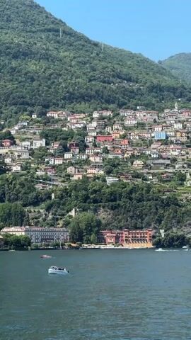 The video starts with a view of a lake with a hillside of houses. The camera then turns to a woman on a boat, who takes a selfie. The woman is wearing a white dress and jewelry. Other people are also on the boat. The weather is sunny.