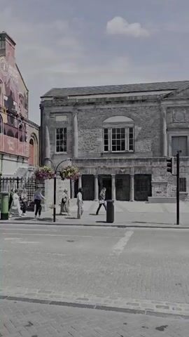 The video pans across a street scene in a town. The buildings are old stone structures, some with multiple stories. In the foreground, there is a brick paved street and sidewalk. A few people are walking on the sidewalk and crossing the street. There are also some benches and planters with flowers. One building has a sign that reads "DINING ROOMS." Another building appears to be a bank. The sky is partly cloudy. The overall impression is a quiet, somewhat dated town center.