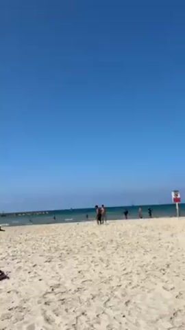 The video shows a sunny beach scene. The sky is clear and blue. The sand is light-colored, and there are footprints visible. Several people are on the beach and in the water. Three shirtless men are playing volleyball near the water's edge. A sign on a post is visible near the right edge of the frame. In the distance, there are small sailboats on the water and a rocky barrier extending into the sea. The overall scene is bright and suggests a pleasant day at the beach.