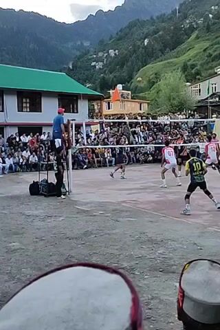 A volleyball game is in progress on an outdoor court. There are many spectators seated and standing around the court, most of them are watching the game. The court is surrounded by a net. Players are on both sides of the net, and they are dressed in athletic attire. One player is in the air, about to hit the ball. The court is surrounded by buildings and a mountain landscape in the background. The weather appears to be overcast.