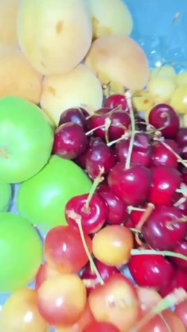 A close-up shot of a decorative glass bowl filled with a variety of fresh fruits. The bowl is situated on a white tablecloth with a subtle floral pattern. The fruits are arranged in distinct sections within the bowl. Starting from the top left and moving clockwise, there are several light-colored, round fruits, likely apricots or peaches. Below these are a cluster of bright green, round fruits, possibly small apples or crabapples. To the right of the green fruits and in the center of the bowl, a large pile of dark red cherries is visible. In front of the dark cherries, there are two smaller piles of cherries: one group of medium red and yellow cherries, and another group of lighter yellow cherries. The overall impression is a vibrant and colorful display of fresh produce. The lighting is soft and diffused, suggesting an indoor setting, possibly a dining room. The camera angle is slightly from above, looking down into the bowl. There are no people or other objects immediately visible in the frame, keeping the focus solely on the fruit arrangement. The movement of the camera is minimal, with slight panning and tilting to capture different angles of the bowl and its contents.