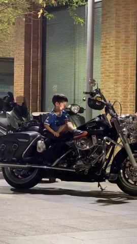 A young child sits on a parked motorcycle. Several people walk by in the background. One person, a young woman wearing glasses, walks past with a bag. Another person, a young man in a red shirt and black shorts, speaks on a phone. The scene takes place outdoors at night.