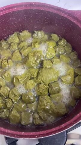 A close-up shot shows a pot of stuffed grape leaves simmering on a stovetop. The grape leaves, which are a deep green color, are tightly packed together and appear to be cooked in a liquid, likely broth or water, as evidenced by the bubbling around them. The pot itself is dark red with a textured exterior and sits on a white stovetop with a black burner grate visible beneath it. The overall scene suggests a home kitchen setting where a meal is being prepared.