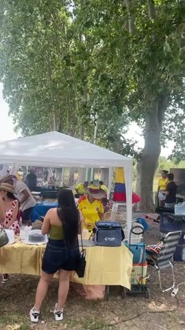 A busy outdoor event, possibly a festival or market, is taking place under a bright, sunny sky with scattered clouds. Tall trees with green foliage line the background, providing shade. Several white tents are set up, some serving as vendor stalls. In the foreground, two young women stand facing away from the camera, one with her arm around the other. They are dressed casually in shorts and t-shirts. Behind them, a yellow-covered table is visible, laden with items including a cake and cups. A woman in a yellow shirt, possibly a vendor, is standing behind the table, interacting with customers. Further in the background, a crowd of people, including adults and children, mill about, some congregating around other tents and stalls. A small child in a yellow shirt runs across the grass in the mid-ground. The ground is covered in dry grass and dirt. The overall atmosphere appears lively and social.