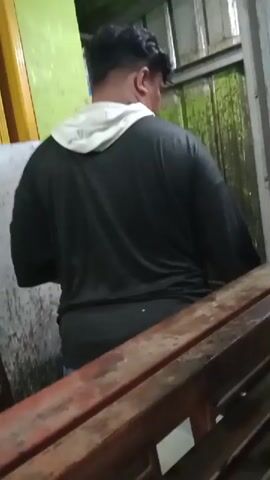 The video shows an adult individual, viewed from behind, standing in what appears to be a utilitarian or semi-enclosed space. The person has dark, short, wavy hair and is wearing a black long-sleeved shirt with a light-colored hood (possibly white or light grey) peeking out from underneath.

The background consists of a green, corrugated metal wall or panel on the right, which shows signs of wear, possibly moss or algae. To the left, there's another lighter-colored wall, possibly yellow or light green. In the foreground, a rusty, dark red metal railing or beam is visible, partially obscuring the lower half of the person.

The individual is largely stationary, facing towards the green metal wall. There's a subtle movement around the 0:02-0:03 mark where the person's right hand slightly moves towards their side, but no clear activity is discernible. The lighting is dim, suggesting it might be evening, night, or an indoor area with limited natural light. No specific location cues beyond the general industrial or storage aesthetic are apparent from the visual information alone. No text is visible in the frame. The overall atmosphere seems mundane.