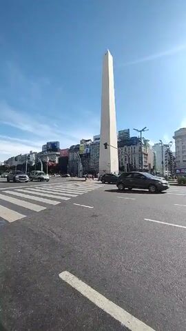 The video shows a low-angle view of the Obelisco in Buenos Aires, Argentina, on a sunny day. Cars are driving on the wide avenue in front of the monument. The street is clearly marked with lanes and pedestrian crosswalks. There are buildings surrounding the Obelisco, some with visible billboards. The sky is blue with some scattered clouds, and the sun is shining brightly, creating lens flare. Pedestrians can be seen near the base of the Obelisco.