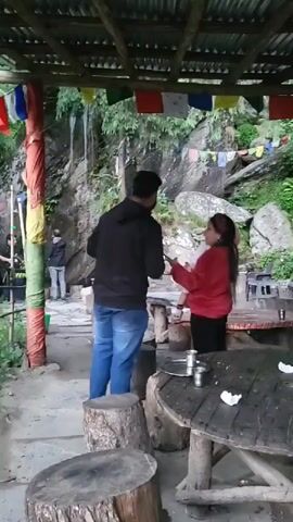 The video shows a partly covered outdoor seating area with several tables made from cut tree trunks. In the background, a rocky cliff is visible with a small waterfall. Prayer flags are hanging above. A woman in a red sweater holds a baby and looks at her phone. A man in a black hooded sweatshirt and blue jeans walks around. The area is decorated with colorful cloth wrapped around a pole and bamboo structures. There are metal cups on the tables. Another person is seen walking in the background.