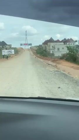 The video shows a view from inside a vehicle traveling on a dirt road. The road is under construction, with loose gravel and visible tire tracks. On the left side of the road, there are several signs and some people standing. Buildings, some appearing residential, are visible on both sides of the road. In the distance, a tall tower with a banner attached to it stands at the crest of a hill. The sky is overcast with dark clouds. A small, white, star-shaped object is visible on the windshield.