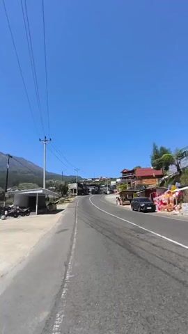 A vertical video shows a paved road curving uphill under a bright blue sky. On the left side of the road, there are a few simple structures, including a white shelter with a motorcycle parked beneath it, and a yellow stall with writing on it, selling goods. Further up the road, a power pole with wires runs overhead. To the left, a large mountain looms in the background, its peak partially obscured by clouds. On the right side of the road, there are a few buildings, including one with a red roof, and a black SUV parked on the side. Trees and vegetation are visible on both sides of the road. The camera appears to be moving forward, possibly from a vehicle or on a bicycle, giving a first-person perspective of the road and its surroundings. There are no people clearly visible in the frame, although there might be some figures in the distance. The overall scene depicts a scenic, possibly rural or mountainous area on a clear day.
