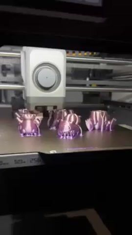 The video shows a 3D printer, a Bambu Lab printer, in operation. Inside the printer enclosure are several small, shiny purple 3D-printed objects resembling cartoonish monster figures, each at varying stages of completion. The printer head moves slowly across the build plate, continuing to lay down material on one of the figures. The build volume of the printer is labeled as 256.