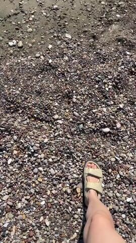 The video opens with a close-up shot of a person's feet wearing beige sandals, walking on a pebbly beach. The camera then pans up to show the shoreline with gentle waves lapping at the sand. Further out, several people are visible in the ocean, swimming and playing in the water. The sky is clear and blue, indicating a sunny day. The overall scene depicts a peaceful and enjoyable day at the beach.