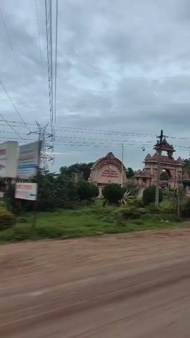 The video shows a view from a moving vehicle passing by a stone gateway, possibly a temple entrance. The gateway is intricately carved with a traditional architectural style. Behind the gateway, there are buildings and trees, including palm trees. In the background, a tall electricity pylon is visible against a cloudy sky. There are power lines crisscrossing the frame. The road in the foreground is unpaved and dusty. In one shot, a few people, seemingly women in traditional Indian attire, walk past the side of the road. The overall impression is of a rural or semi-rural setting in India.