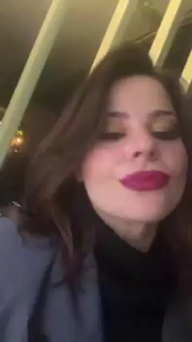 A woman with brown hair and red lipstick is the main subject of this close-up video. She is wearing a black turtleneck and a grey blazer. The camera angle is low, looking up at her face. The woman sticks her tongue out and makes exaggerated mouth movements, as if mimicking speaking or singing without making audible sounds. The background is blurry and appears to be an outdoor environment at night, with faint lights visible.