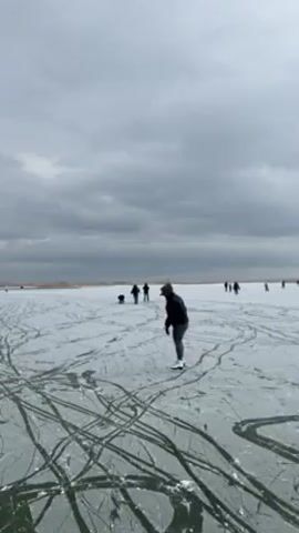 The video shows a woman ice skating on a frozen lake or large body of water. The ice is covered in skate marks.  Other people are visible skating or walking in the distance.  The sky is overcast. The woman is wearing dark clothing and white skates.  She starts off looking down, then looks up and smiles.  Another person skates near her.