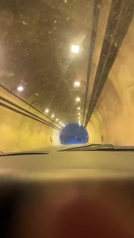 A car drives through a well-lit tunnel, exiting to a scene of wooded mountains under a partly cloudy sky. The camera's perspective is from inside the car, looking through a windshield. The tunnel interior has yellow walls and a series of lights overhead. Outside, the landscape features a mountain covered with trees, street lamps, and signage. The sky above is a blend of blue and cloudy white.
