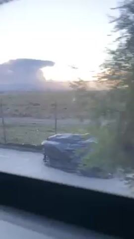 The video, shot from inside a moving vehicle, shows a view of an open, grassy landscape under a bright, clouded sky, suggesting either sunrise or sunset. The camera moves along a road, showing glimpses of the terrain, roadside structures, and a few pedestrians walking along the pavement. The footage captures the ambiance of an open space with the interplay of natural light and sparse human presence.